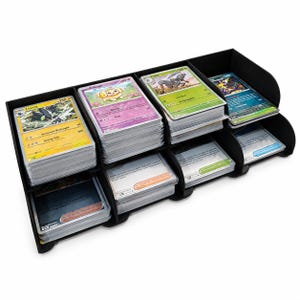 May include: A black plastic storage organiser with eight distinct compartments, each filled with stacks of trading cards. The cards feature various colourful illustrations, character names, and text, organised in two rows within the structure.