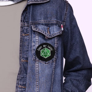 May include: A black and green embroidered patch with a white outline. The patch features a green 20-sided die with a white "1" on it. The text "Just My Luck!" is written in white around the die.
