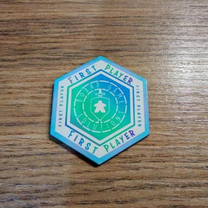 May include: A hexagonal game token with a gradient of blue and green. The token reads "FIRST PLAYER" around the edges and "BOARD GAME SOLUTIONS" in the center, with a stylized person icon. The token is on a wooden surface.