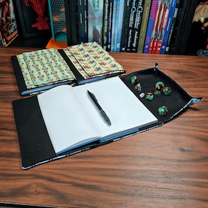 Puede incluir: Cuadernos con cubiertas estampadas, uno abierto con un bolígrafo y una bandeja para dados. Las cubiertas presentan diseños con frascos y espadas sobre un fondo verde claro y amarillo. El cuaderno abierto tiene páginas rayadas.