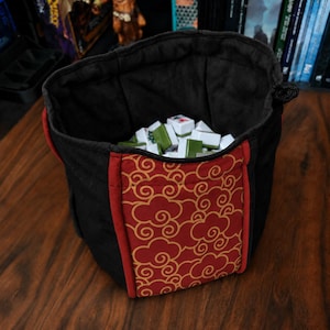 May include: A black fabric drawstring bag with a red and gold cloud pattern panel on the front. The interior is lined with black fabric and contains a collection of white and green rectangular mahjong tiles with various markings.