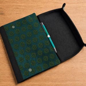 Könnte beinhalten: Ein dunkelblaugrünes Falt-Tablett mit einem sich wiederholenden geometrischen Sternmuster in Grün. Das Tablett verfügt über schwarze Seitenteile, silberne Druckknöpfe und einen blaugrünen Kugelschreiber, der diagonal auf der Innenfläche liegt.