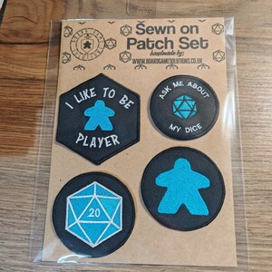 May include: A pack of four black and blue gaming patches. One patch reads "I LIKE TO BE PLAYER", another says "ASK ME ABOUT MY DICE". The set includes a 20-sided die and a player piece. Made by www.boardgamesolutions.co.uk.