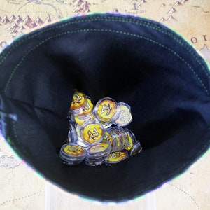 以下が含まれることがあります： 緑と白のパターンがついた縁取りのある黒い布製の袋。袋には、黄色と黒のデザインが施された透明なプラスチック製のトークンが入っています。トークンには「1」という数字が印刷されています。