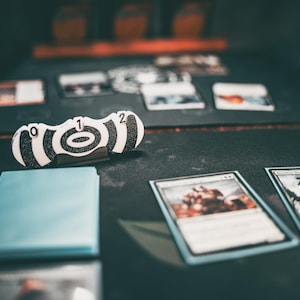 以下が含まれることがあります： 黒と白のターゲットカウンターに、0、1、2の数字が印刷されています。カウンターは、背景にトランプがある黒い表面の上に置かれています。