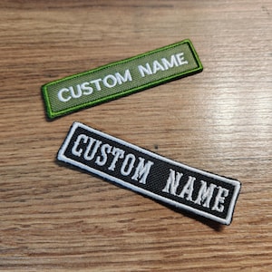 Puede incluir: Dos parches rectangulares bordados con bordes cosidos en blanco. Un parche es de color verde oliva con el texto blanco CUSTOM NAME, y el otro es negro con el texto blanco CUSTOM NAME. Ambos presentan un estilo de fuente limpio y uniforme.