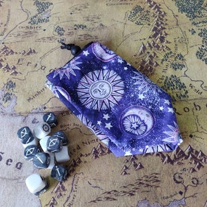 May include: A purple dice bag with a celestial design featuring suns, moons, and stars. The bag is closed with a black drawstring. A set of dice with white and gray markings are next to the bag. The background is a map.