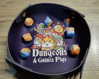 Puede incluir: Una bandeja de dados de cuero negro con una ilustración colorida de conejillos de indias vestidos de aventureros. La bandeja tiene el texto "Dungeons & Guinea Pigs" impreso en ella. Hay seis dados coloridos dentro de la bandeja.