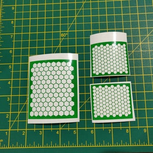 May include: Three white vinyl stickers with a green border, each featuring a pattern of white hexagons on a green background. The stickers are on a green cutting mat with inch markings.