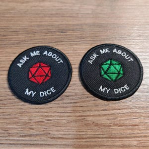 Op de afbeelding: Twee zwarte, ronde patches met witte tekst: "ASK ME ABOUT MY DICE". Eén heeft een rode polyhedrale dobbelsteen, de andere een groene. De patches zijn waarschijnlijk voor een gamer.