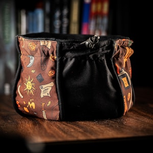 May include: A brown and black drawstring bag with a black lining. The bag is decorated with a pattern of various fantasy and role-playing game symbols, including dice, swords, and scrolls.