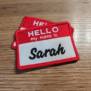 May include: Two red and white embroidered name tags. The top tag reads "HELLO my name is Sarah" in black script. The tags have a red border and are on a wooden surface.