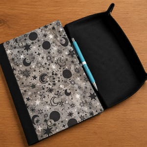 May include: A rectangular black tray with a folding flap featuring a gray and black celestial pattern of moons and stars. The tray includes two silver metal snaps on the flap and a bright blue retractable ballpoint pen resting inside.