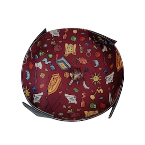 May include: A red felt dice tray with a black border and a variety of colorful illustrations, including dice, scrolls, and a sun. The tray is designed for tabletop role-playing games.