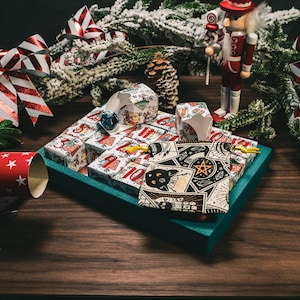 Könnte beinhalten: Eine türkisfarbene Schachtel mit einem Adventskalender für Weihnachten mit einem schwarzen Kater und Tarotkarten-Designs. Die Schachtel ist von festlichen Dekorationen umgeben, darunter rot-weiß gestreifte Schleifen, eine Nussknackerfigur und ein Weihnachtscracker.