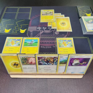 May include: A close-up of a Pokemon trading card game setup with a variety of cards on a black playmat. The cards are arranged in a grid pattern, with a deck of cards in the upper right corner. The cards feature various Pokemon characters, including Pikachu, Charmander, and Bulbasaur. The cards are all in good condition and have a glossy finish.