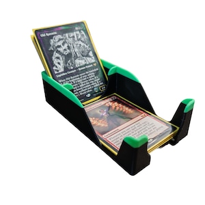 Puede incluir: Soporte para tarjetas negro y verde con dos compartimentos. El soporte está diseñado para guardar tarjetas coleccionables. El compartimento superior contiene una tarjeta con el texto "Old Rutstein". El compartimento inferior contiene una tarjeta con el texto "Enchantment".