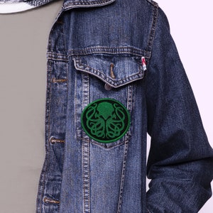 May include: A green and black embroidered patch of a stylized octopus with tentacles, in a circle, sewn onto a blue denim jacket.