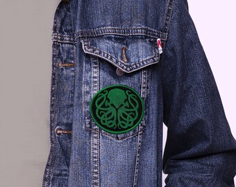 May include: A green and black embroidered patch of a stylized octopus with tentacles, in a circle, sewn onto a blue denim jacket.