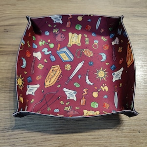 May include: A red felt dice tray with a black border. The tray is decorated with a repeating pattern of fantasy-themed illustrations, including dice, swords, scrolls, and other magical items.