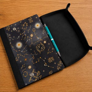 May include: A black rectangular valet tray with a folding flap featuring a gold celestial print of constellations, suns, and zodiac symbols. The tray has silver metal snap buttons at the corners and contains a single teal and silver ballpoint pen.