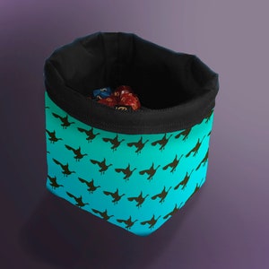May include: A turquoise fabric dice bag with a black drawstring and a black trim. The bag is decorated with a repeating pattern of black flying birds. The bag is filled with colorful dice.