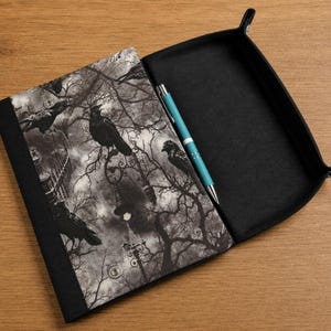 May include: A black fabric valet tray with a rectangular shape and snap-button corners. The interior features a monochromatic print of crows perched on bare tree branches and street lamps. A bright turquoise ballpoint pen rests inside.