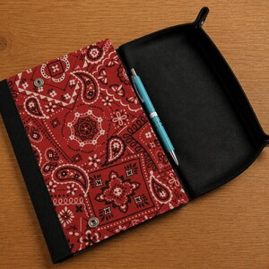 May include: A rectangular valet tray featuring a red and white paisley bandana print fabric on the exterior flap and a solid black interior. The tray includes a silver metal snap button closure and a light blue ballpoint pen resting inside.