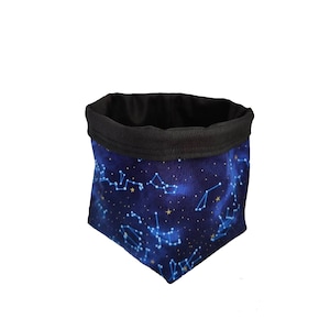 Puede incluir: Una cesta de almacenamiento de tela con un diseño celestial. La cesta presenta un fondo azul oscuro con constelaciones y estrellas doradas. El interior y el borde superior son negros.