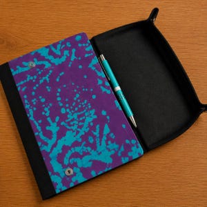 May include: A rectangular black valet tray with a purple and turquoise splatter-patterned cover secured by two silver metal snaps. The tray features a black interior lining and includes a turquoise ballpoint pen resting on the surface.