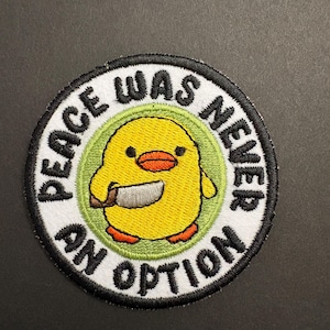 May include: Round embroidered patch featuring a yellow chick holding a knife. The chick is inside a green circle, surrounded by a white border with the text "PEACE WAS NEVER AN OPTION" in black.