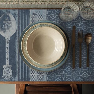 May include: A table setting with a blue and white patterned placemat, a beige bowl, a blue plate, and a smaller plate with a green trim. The setting includes a knife, fork, and spoon with a bronze finish, and two clear glasses.