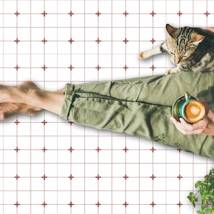 Op de afbeelding: Een persoon zit op een witte betegelde vloer met rode kruismarkeringen. Ze houden een kopje koffie vast en een kat zit op hun schoot.