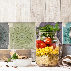 Może przedstawiać: Szklany słoik wypełniony sałatką z quinoa, kukurydzą, groszkiem i pomidorami cherry. Słoik stoi na białym drewnianym stole z miską pieprzu, drewnianą łyżką i zielonym liściem. Tło stanowi ściana z czterema ozdobnymi płytkami z wzorami mandali.