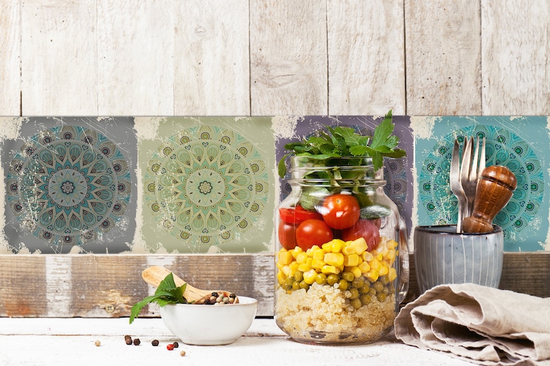 Może przedstawiać: Szklany słoik wypełniony sałatką z quinoa, kukurydzą, groszkiem i pomidorami cherry. Słoik stoi na białym drewnianym stole z miską pieprzu, drewnianą łyżką i zielonym liściem. Tło stanowi ściana z czterema ozdobnymi płytkami z wzorami mandali.