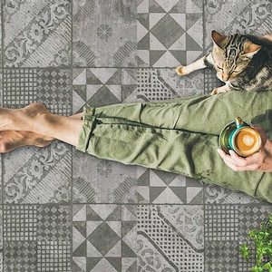 以下が含まれることがあります： 緑色のズボンを履いた人が、模様のあるタイル張りの床に座っており、猫が膝の上に乗っています。右手にコーヒーカップを持っています。