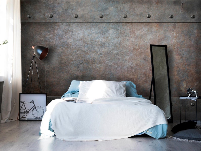 May include: A white bed with a blue blanket and white pillows sits in a room with a gray concrete wall. The wall has a row of silver rivets along the top. A black floor lamp with a black shade is in the corner of the room. A black framed picture of a bicycle is on the floor next to the bed.