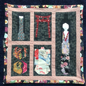 May include: A quilted wall hanging with a Japanese theme. It features a red torii gate, a kimono, a traditional Japanese doll, and a geometric pattern.