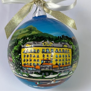 May include: Hand-painted glass Christmas ornament featuring a yellow building with a blue sky and green mountain backdrop. The ornament is tied with a gold and white ribbon. The building is reflected in the water below.