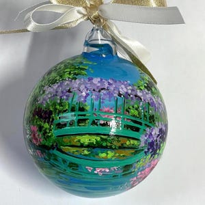Può includere: Un ornamento sferico in vetro con una scena dipinta a mano raffigurante un ponte ad arco verde sopra uno stagno blu con glicini viola e vegetazione rigogliosa. L'ornamento è sormontato da un nastro dorato e bianco legato a fiocco.