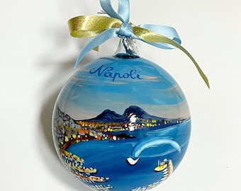 May include: A round glass ornament featuring a hand-painted coastal scene of Naples, Italy, with the word Napoli written in blue script at the top. The ornament is topped with a light blue satin ribbon tied in a bow for hanging purposes.
