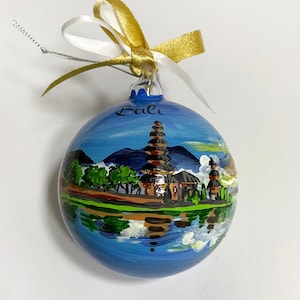 Peut inclure: Une boule de Noël en verre ronde ornée d'un paysage peint à la main représentant un temple au bord d'un lac avec le mot Bali écrit en haut. La décoration est surmontée d'un nœud en ruban doré et blanc et d'une fine ficelle argentée.