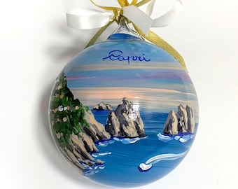 May include: A spherical glass ornament featuring a hand-painted coastal scene of Capri with rocky cliffs, blue water, and a small boat. The word Capri is written in blue script at the top. A gold and white ribbon is tied in a bow at the top.