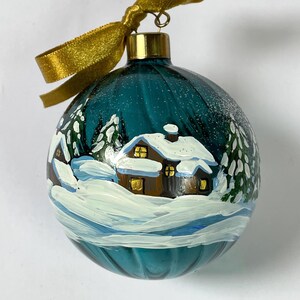 May include: A spherical teal glass ornament featuring a hand-painted winter scene with brown cabins, snow-covered roofs, and evergreen trees. The ornament is topped with a gold metal cap, a gold wire hook, and a tied gold ribbon bow at the top.