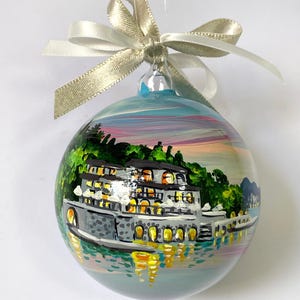 May include: A spherical glass ornament featuring a hand-painted scene of a multi-story building with illuminated windows situated by a body of water, surrounded by green trees and a sunset sky, topped with a shimmering champagne-colored ribbon bow.