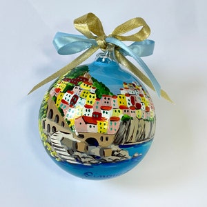 Può includere: Un addobbo natalizio in vetro dipinto a mano che raffigura un pittoresco villaggio italiano con cielo blu e mare. L'addobbo è legato con un nastro blu e oro.