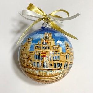 Può includere: Un addobbo natalizio dipinto a mano che raffigura un edificio con una torre dell'orologio, una bandiera spagnola e uno sfondo di cielo blu.
