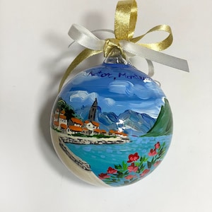 May include: A round glass ornament featuring a hand-painted coastal scene of Kotor, Montenegro, with blue water, mountains, and red-roofed buildings. The ornament is topped with a gold and white ribbon bow for hanging, and the text Kotor, Montenegro is painted on the upper portion.