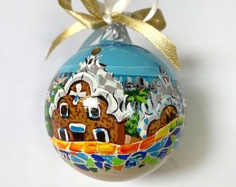 May include: A round glass Christmas ornament featuring a hand-painted scene of a brown building with a white roof, a blue sky, and a colorful mosaic base. The ornament is topped with a shiny gold ribbon tied into a bow for hanging purposes.