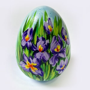 Può includere: Un uovo dipinto a mano raffigurante una vivace scena floreale con iris viola e centri giallo brillante. I fiori sono circondati da lunghe e sottili foglie verdi su uno sfondo azzurro, creando un dettagliato disegno botanico.